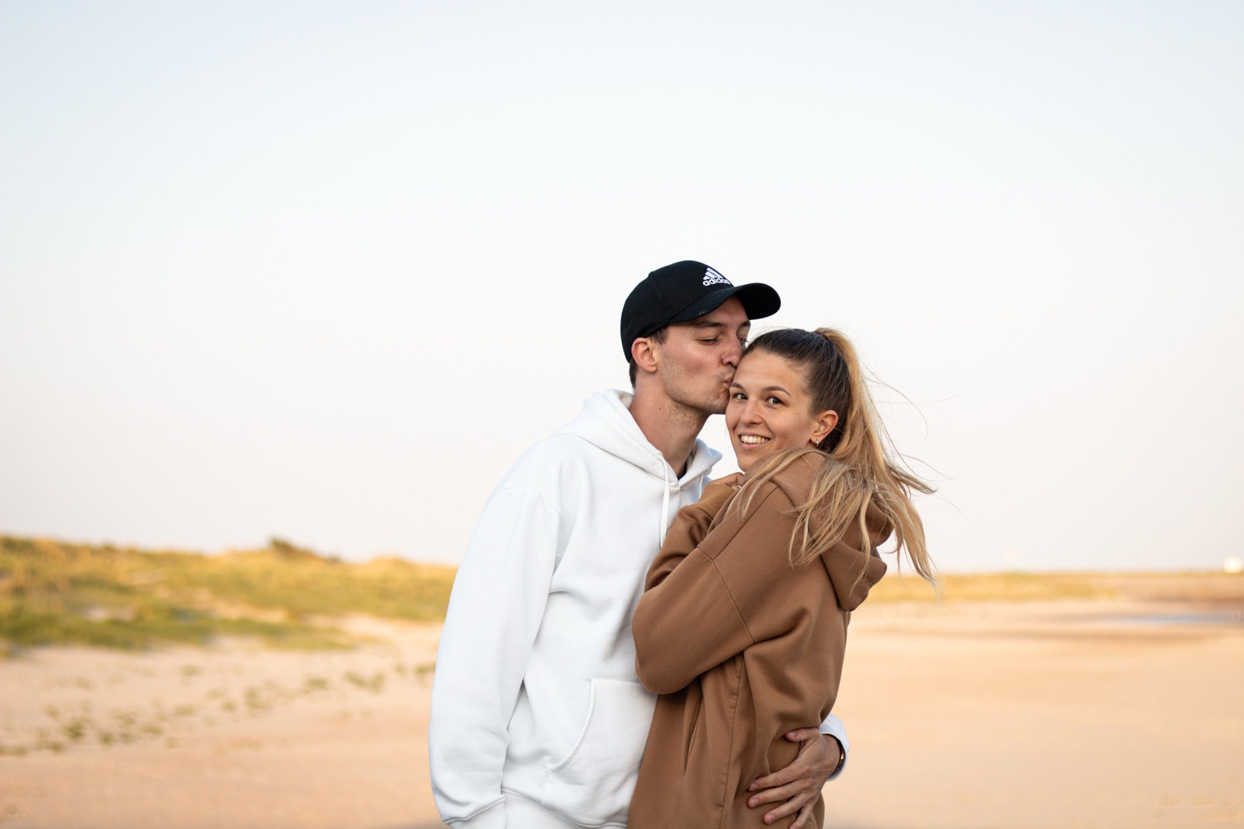 Alex küsst Vicky auf die Schläfe, beide eng umarmt am Strand, dahinter Dünen und ein heller Abendhimmel