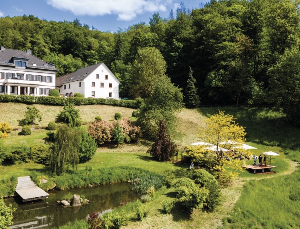 Das Hofgut Hohenstein von oben: weiße Gutshäuser am Waldrand, davor eine Wiese mit Teich und Steg